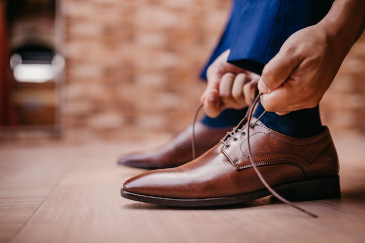 A Person Tying The Lace Of A Brown Leather Shoe