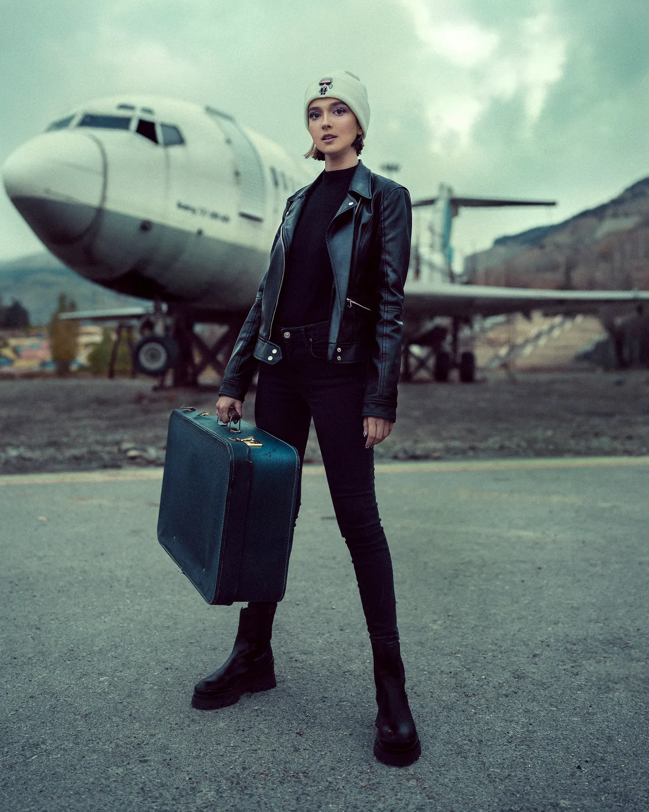all-black-airport-outfit Woman in an all-black airport outfit with leather jacket, beanie, and suitcase