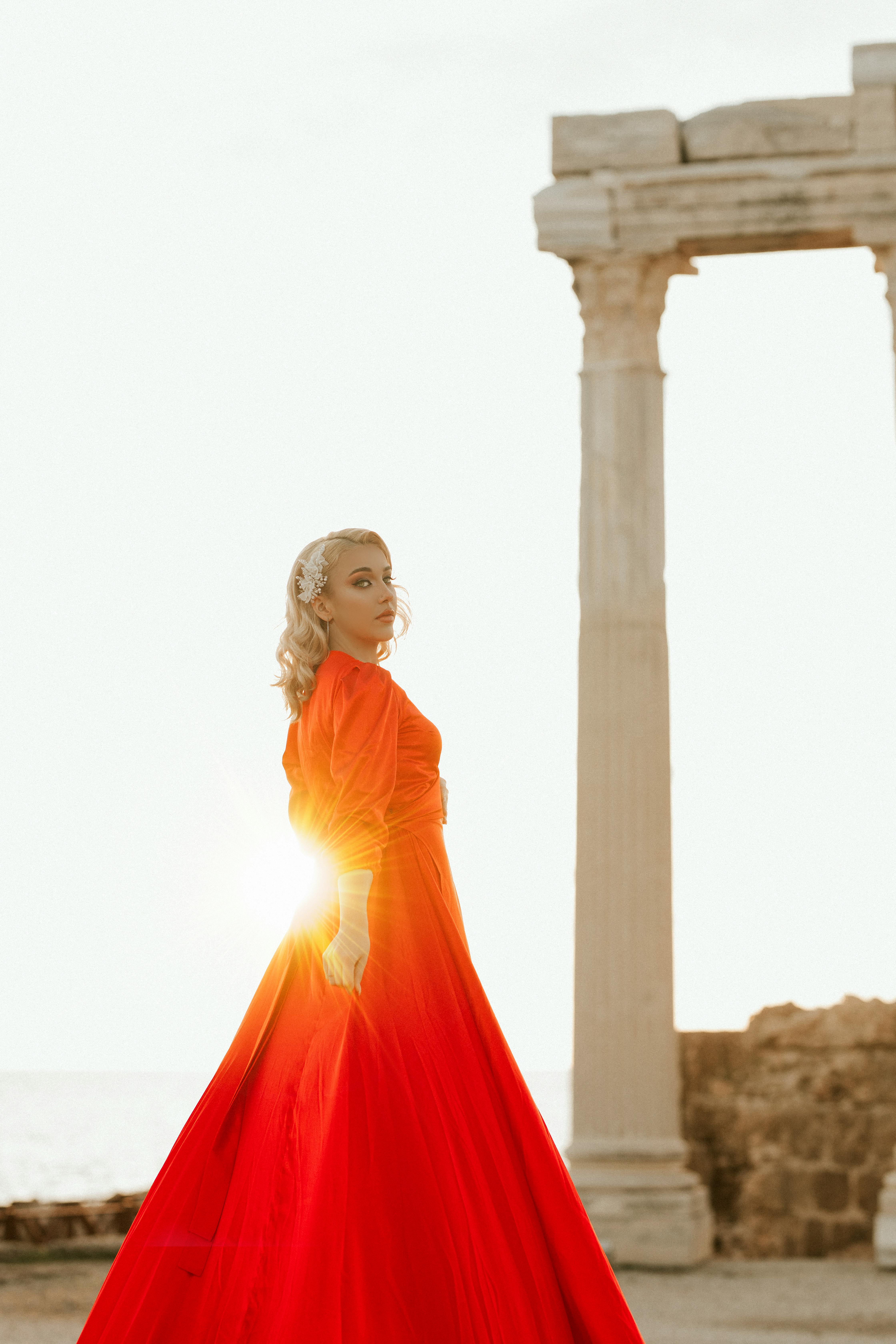 A stylish woman in a flowing red dress poses by ancient pillars at sunset.