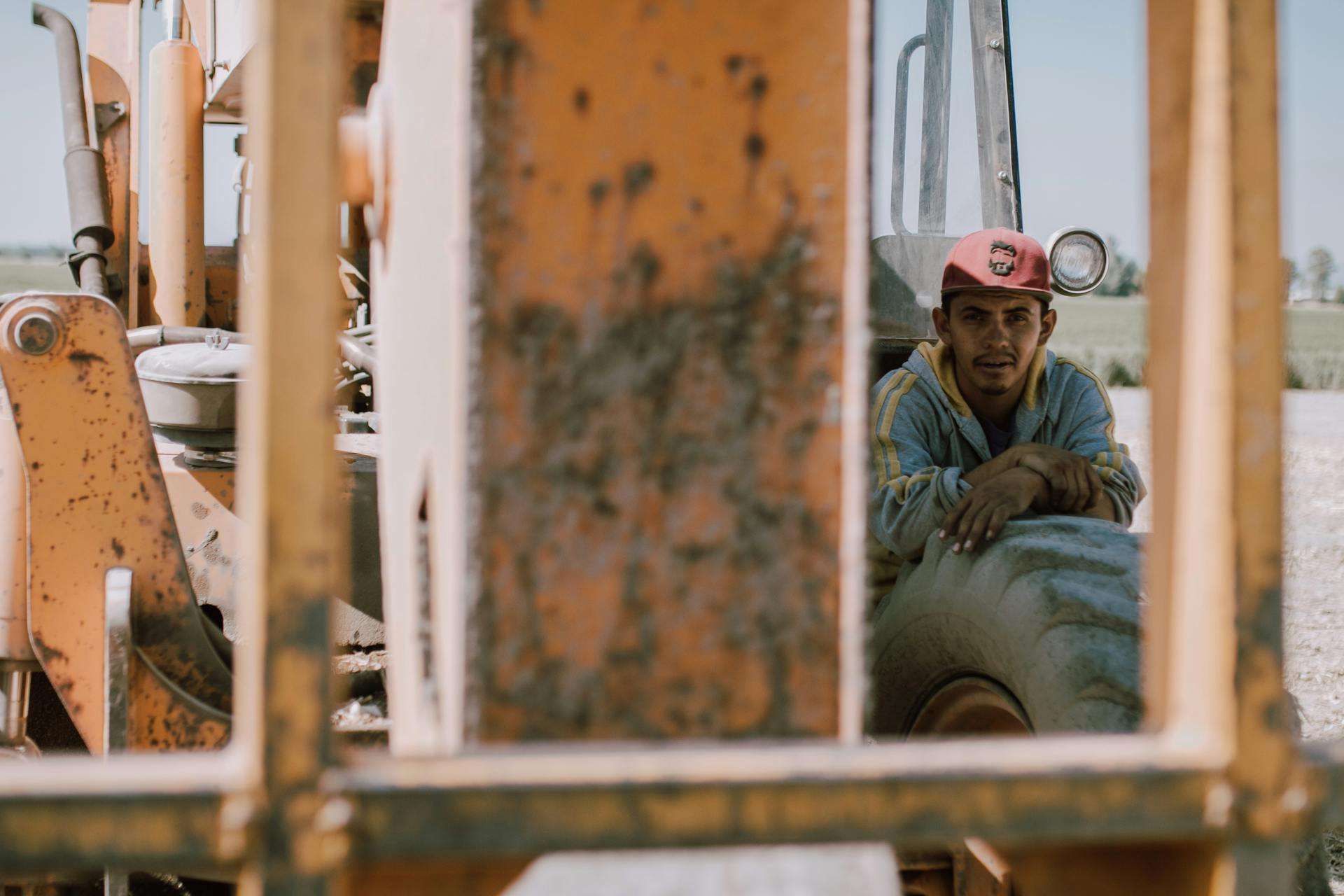 A construction worker in a cap leans on an excavator tire during a sunny day.