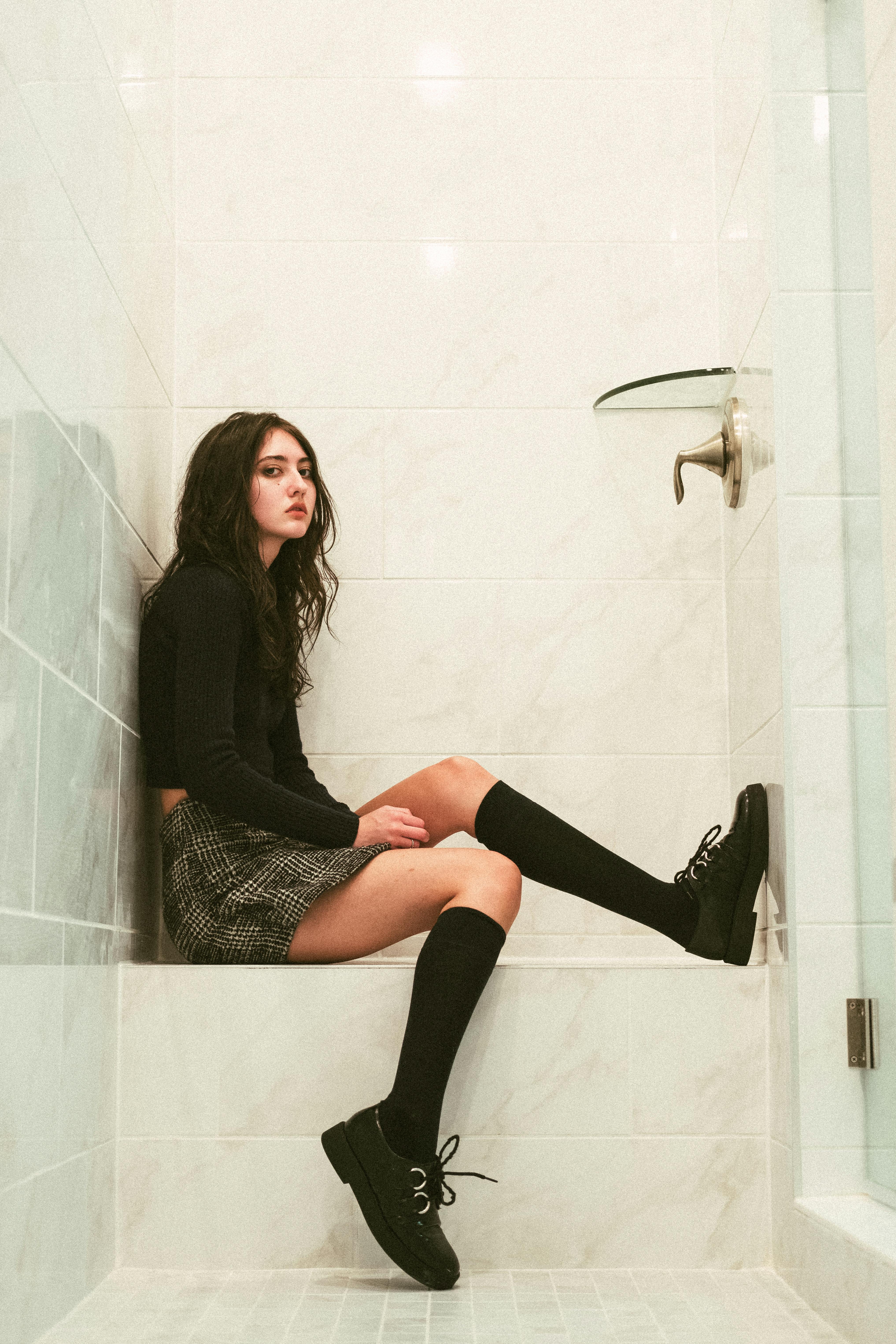 Stylish teenager in monochromatic outfit sitting in a sleek, minimalist bathroom.