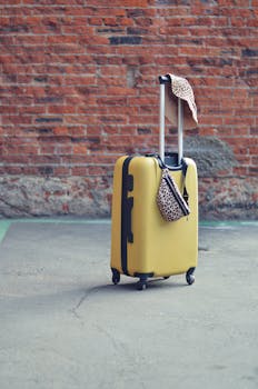 A yellow suitcase with a hat and bag against a brick wall, symbolizing travel and adventure.