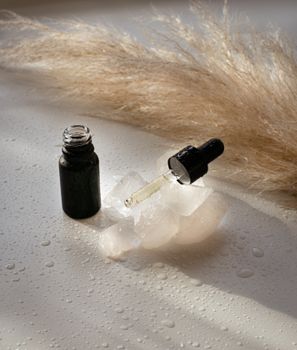 Close-up of a serum bottle and pipette on a wet surface with dried grass in soft lighting.