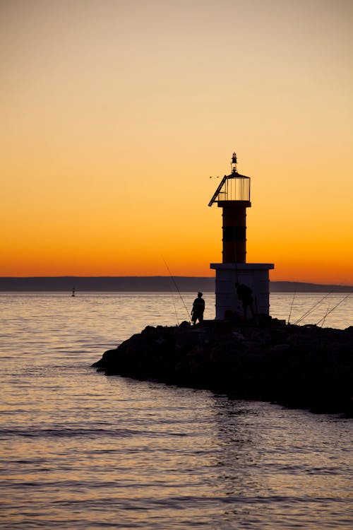 A Silhouette of a Person Fishing by a Lighthouse during Sunset