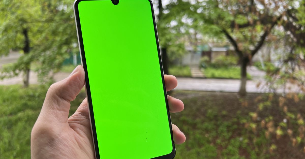 A hand holding a smartphone with a broken screen, nature background behind it