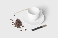 Minimalist flat lay of coffee beans, empty cup, saucer, stirrer, and sachet on white background.