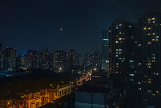 A stunning cityscape under a night sky with a crescent moon, showcasing urban lights and skyscrapers.