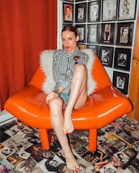 Woman sitting barefoot on an orange chair with a striped shirt in a vintage setting.