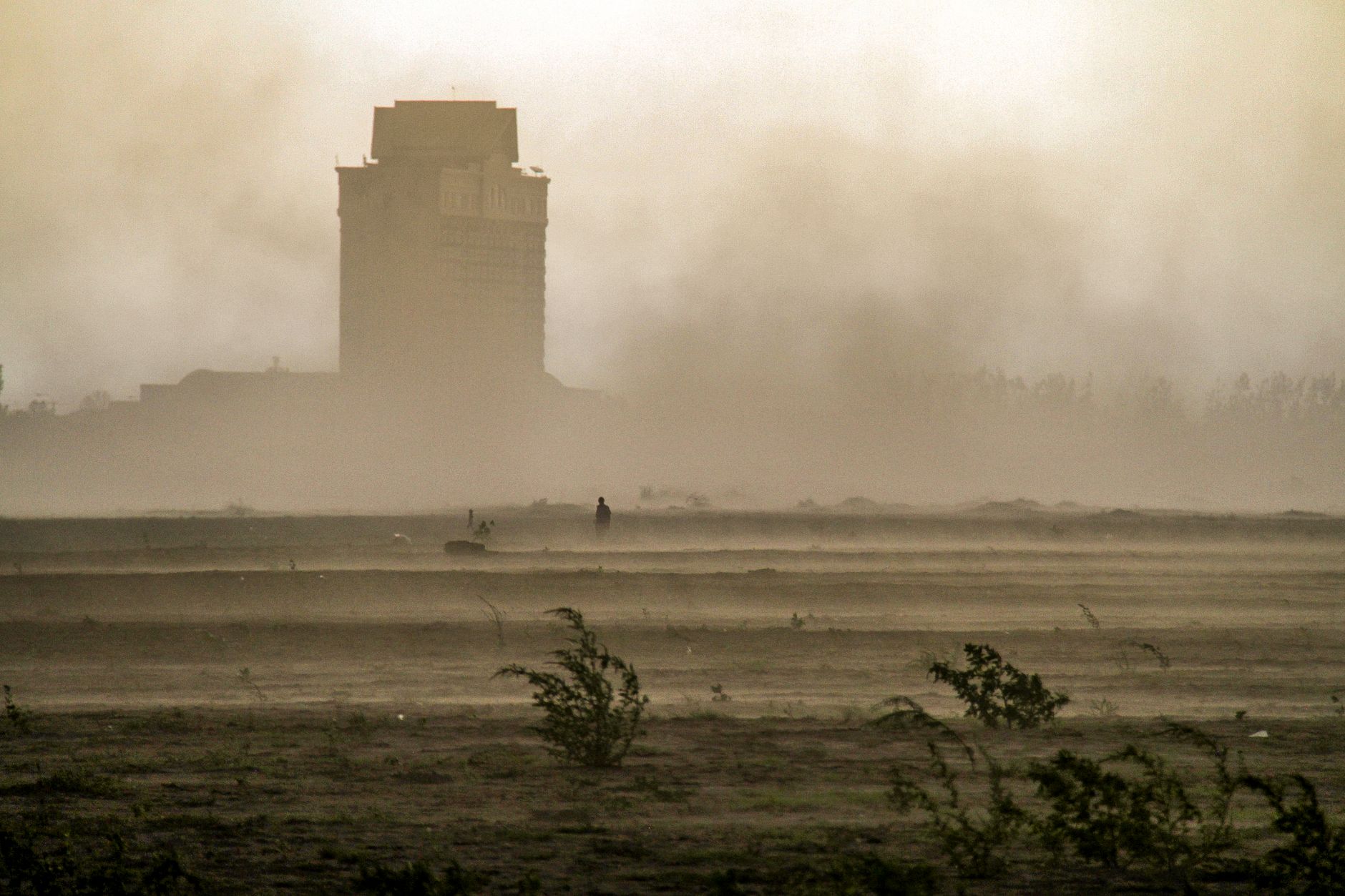 A dramatic scene of a dust storm engulfing a building and sparse vegetation in the foreground.