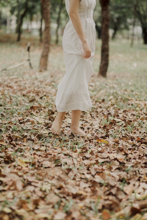 A Barefooted Person in White Dress Standing on the Grassy Ground Full of Dried Leaves