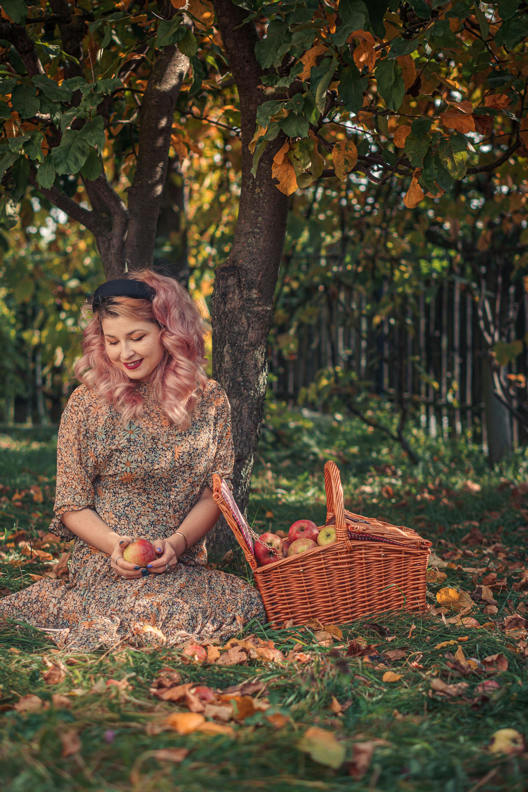 Woman with pink hair sits with apples and a basket under a tree in autumn.