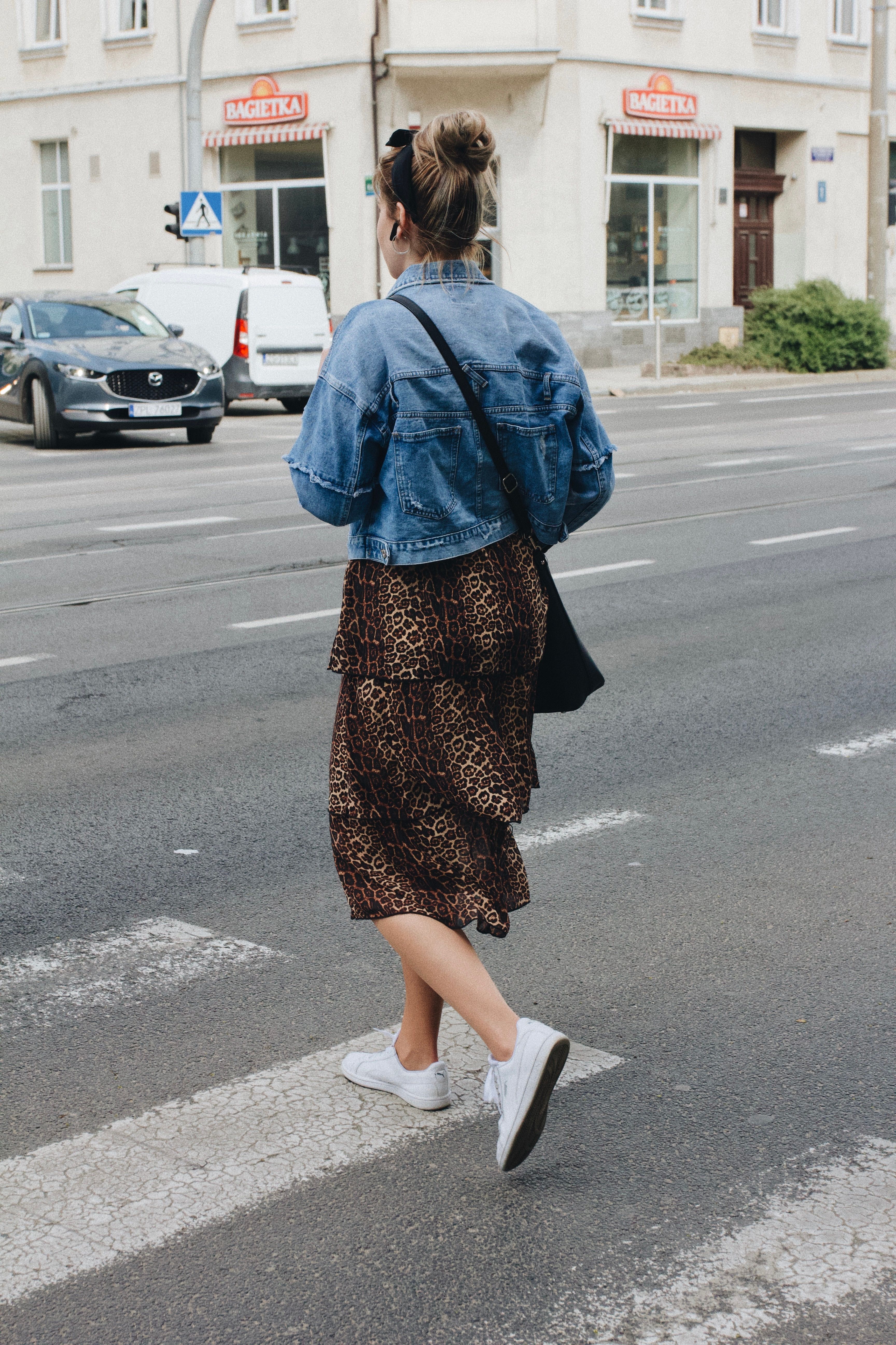 Woman in a denim jacket with a leopard midi skirt and white sneakers crossing the street