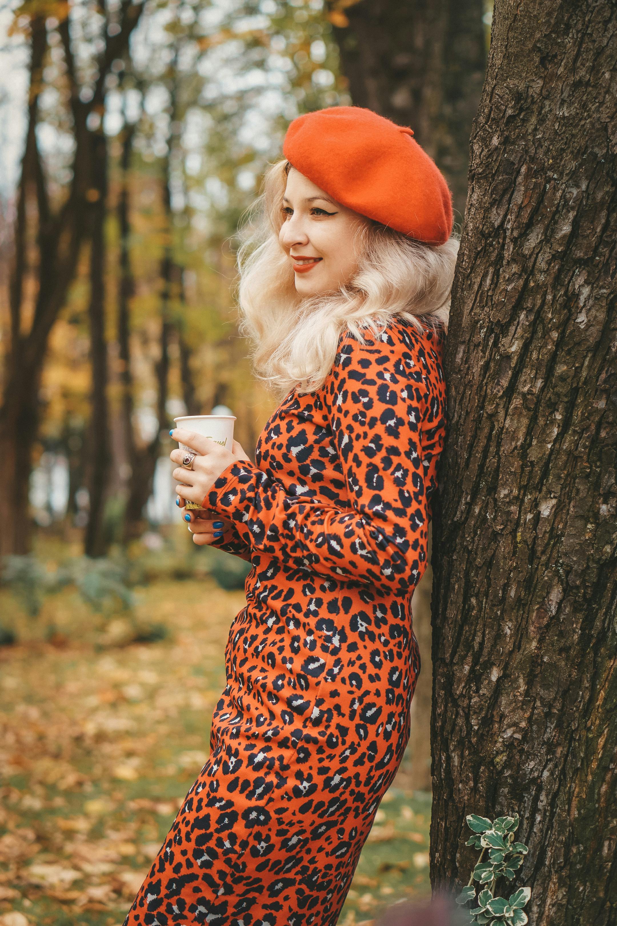 Stylish woman in an autumn park wearing a red beret and leopard print dress, holding coffee.