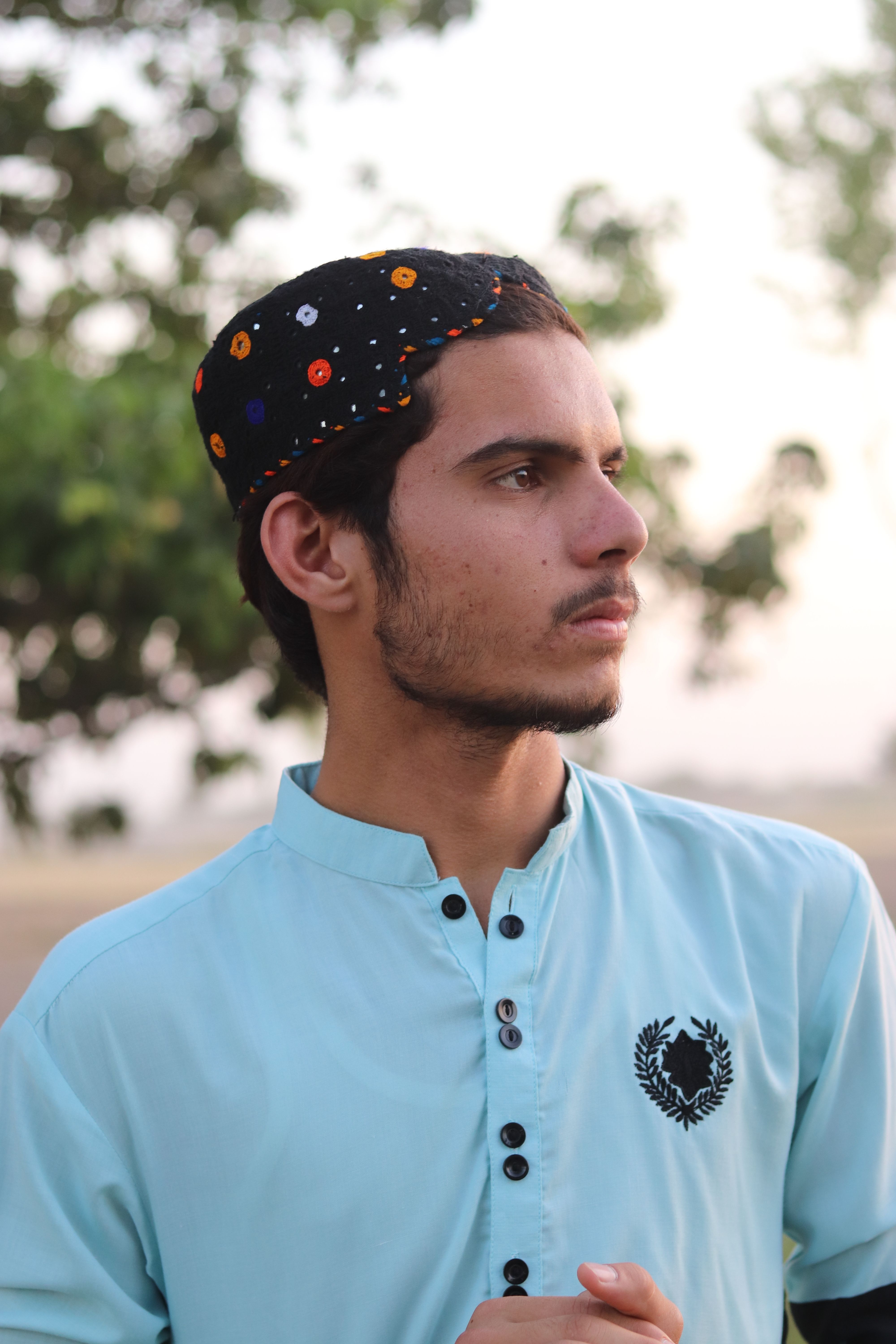 A young man in traditional attire with colorful cap, looking thoughtfully outdoors.