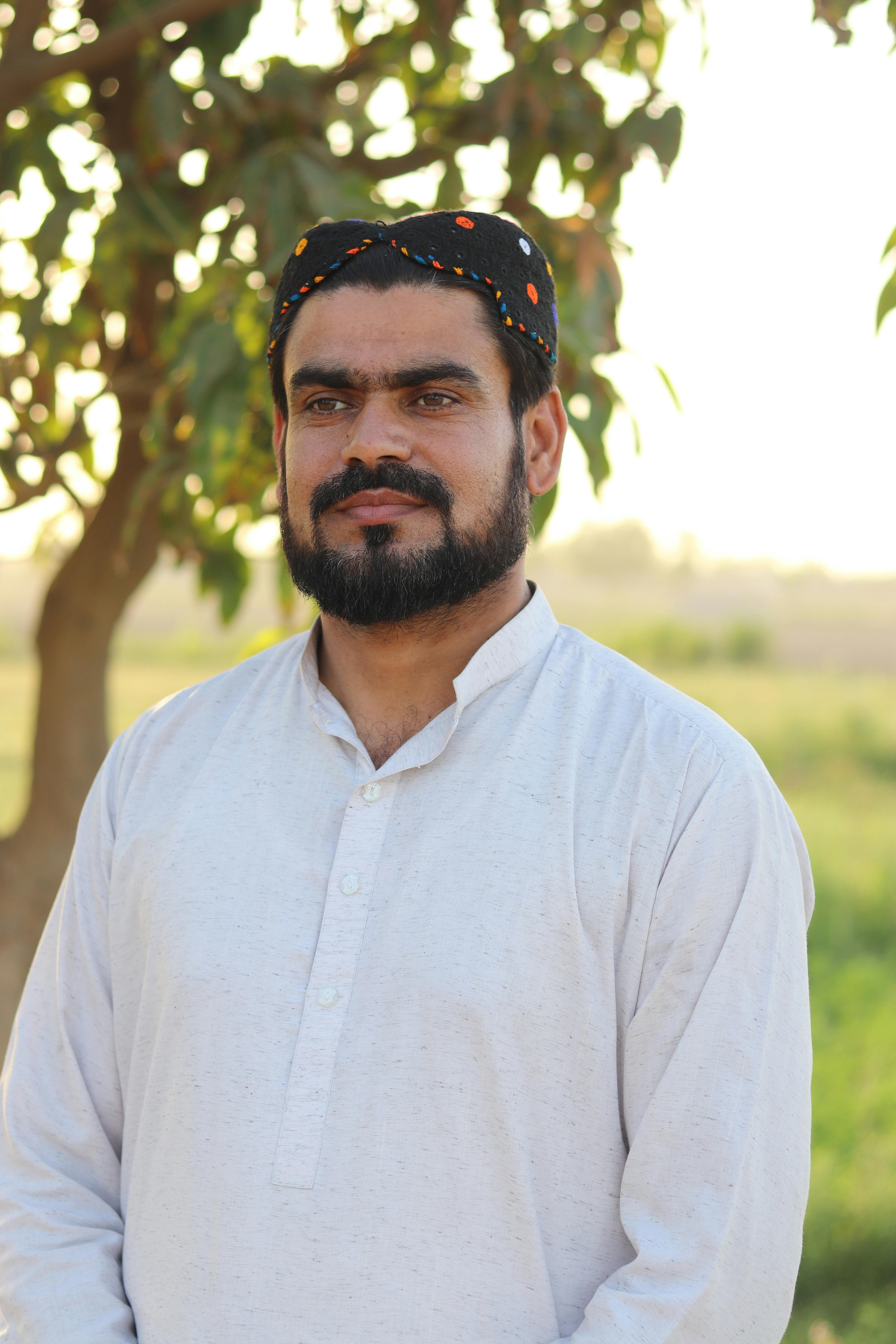 A confident man with a mustache and beard wearing traditional attire and standing outdoors near a tree.