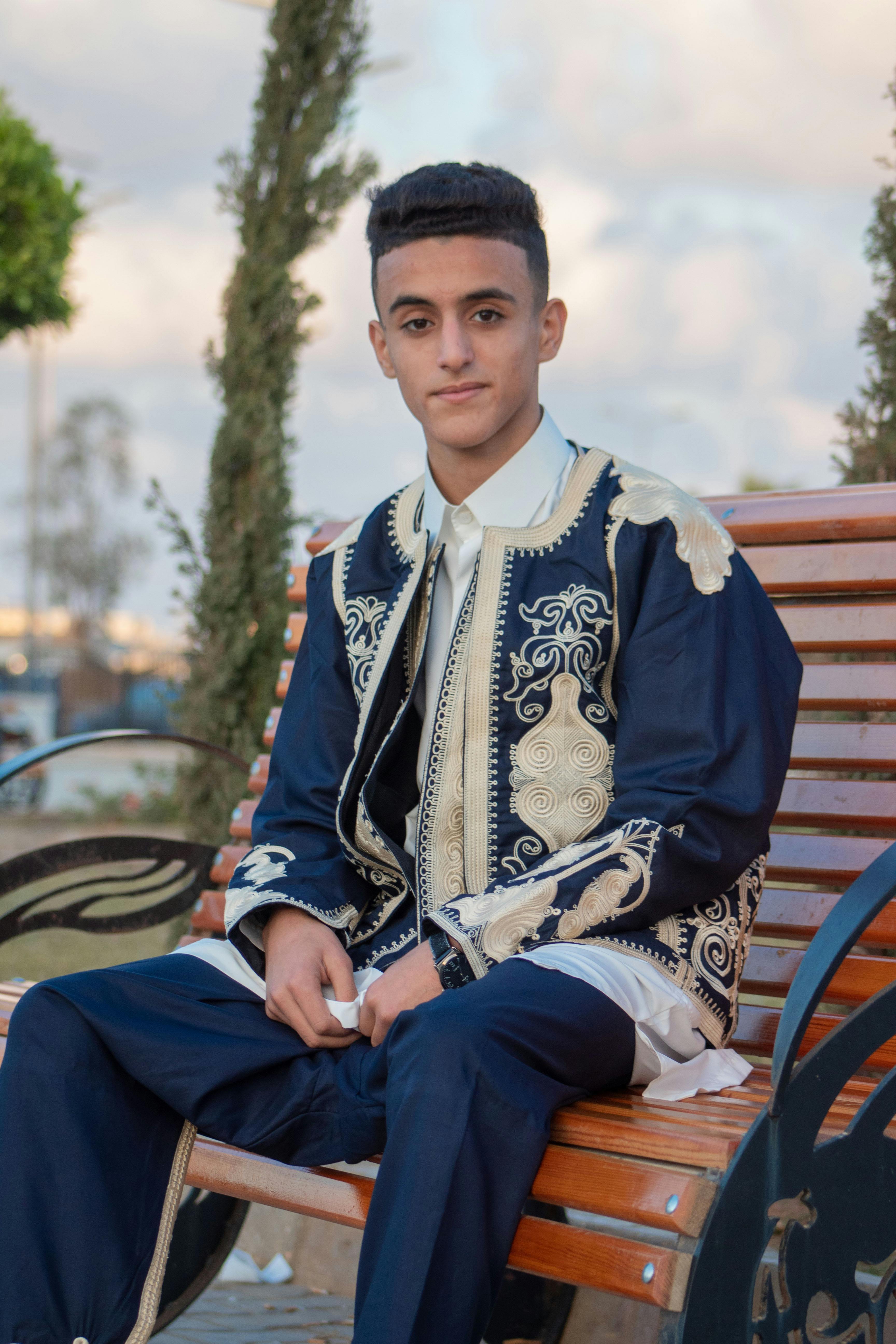 Teenager in traditional embroidered clothing sitting on a wooden bench outdoors, showcasing cultural heritage.