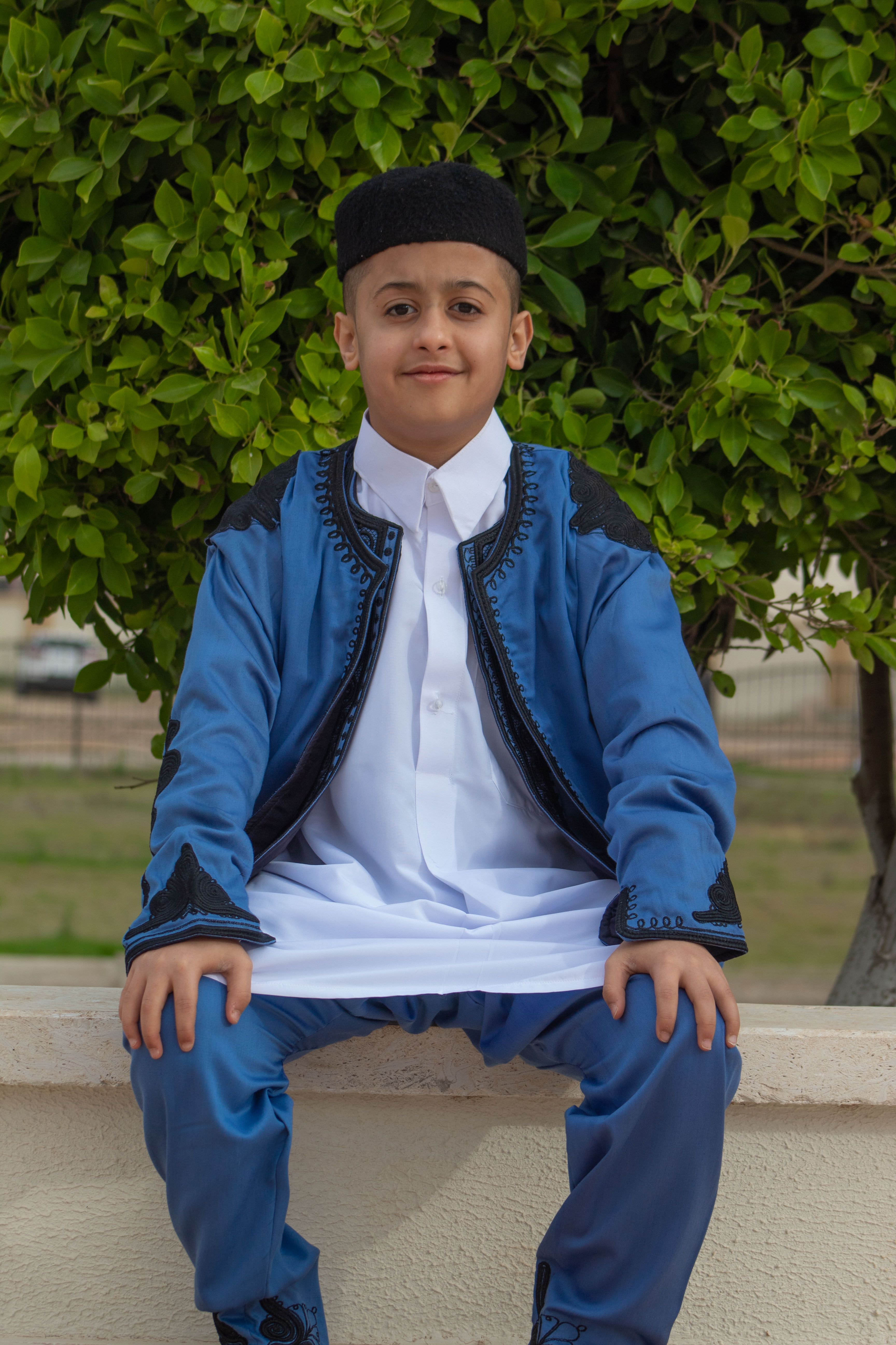 A cheerful young boy wearing traditional blue clothing sits outside against a green leafy backdrop.