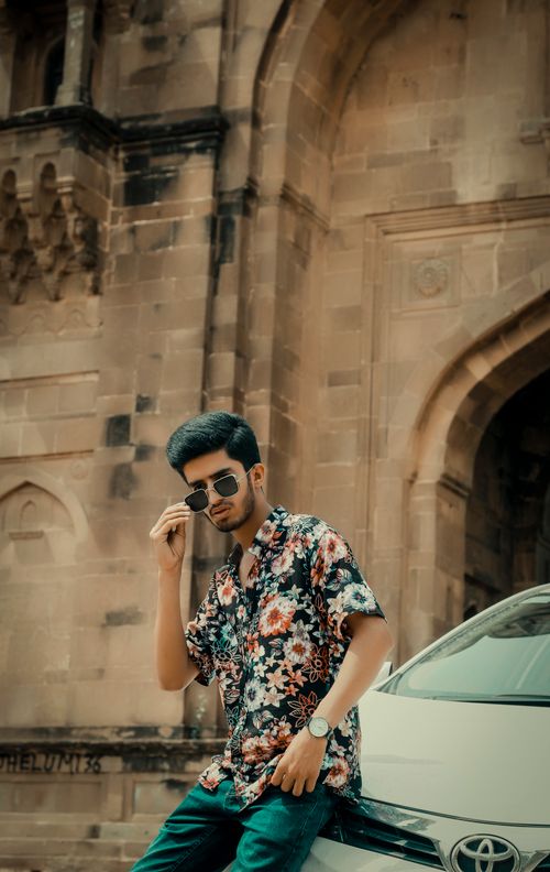 Free A fashionable young man in a floral shirt posing with sunglasses by a historical building and a car. Stock Photo