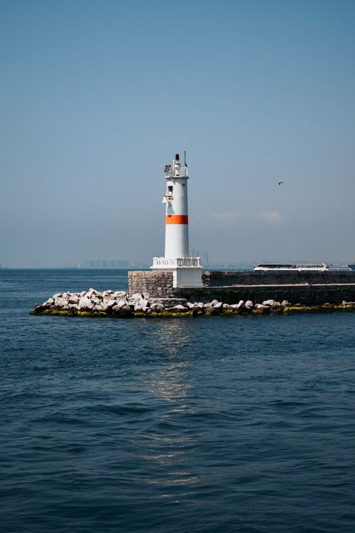 Free A solitary lighthouse stands against a clear blue sky by the seashore, guiding nautical travelers. Stock Photo