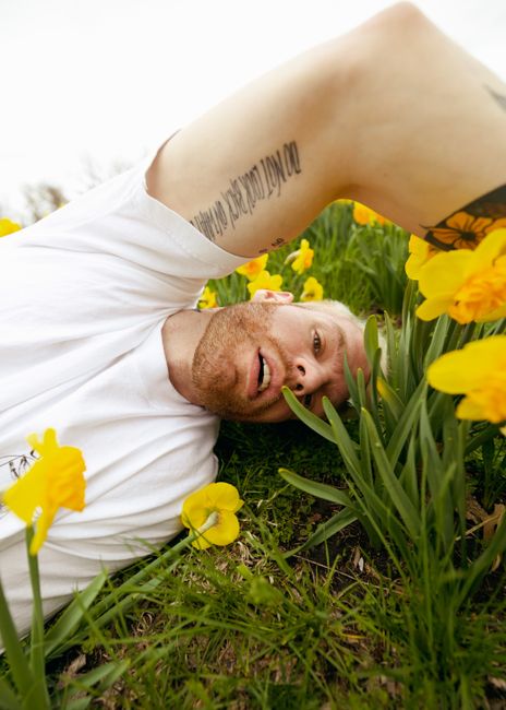 Young man posing in a field of yellow flowers, wearing a white shirt, during a sunny summer day.