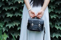 一名女子身穿条纹连衣裙，手持黑色皮革手提包，背景是郁郁葱葱的绿叶