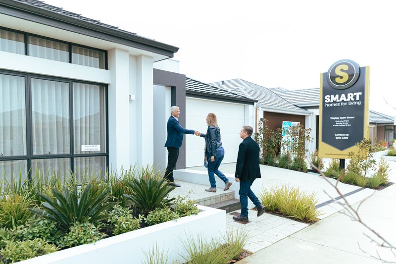 A realtor shakes hands with potential buyers outside a modern model home.