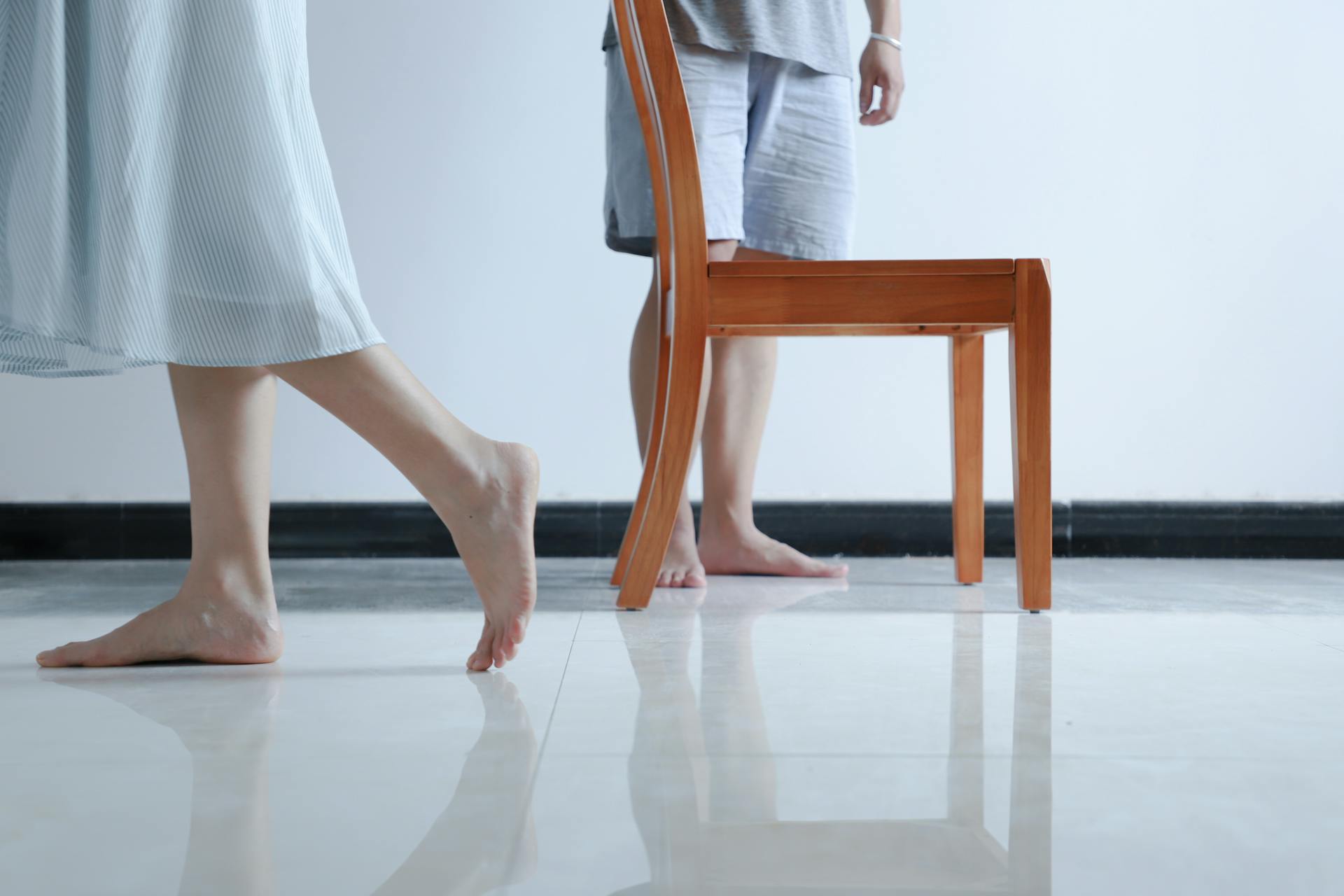 Two people walk barefoot on a sleek polished floor beside a wooden chair in a minimalist room.