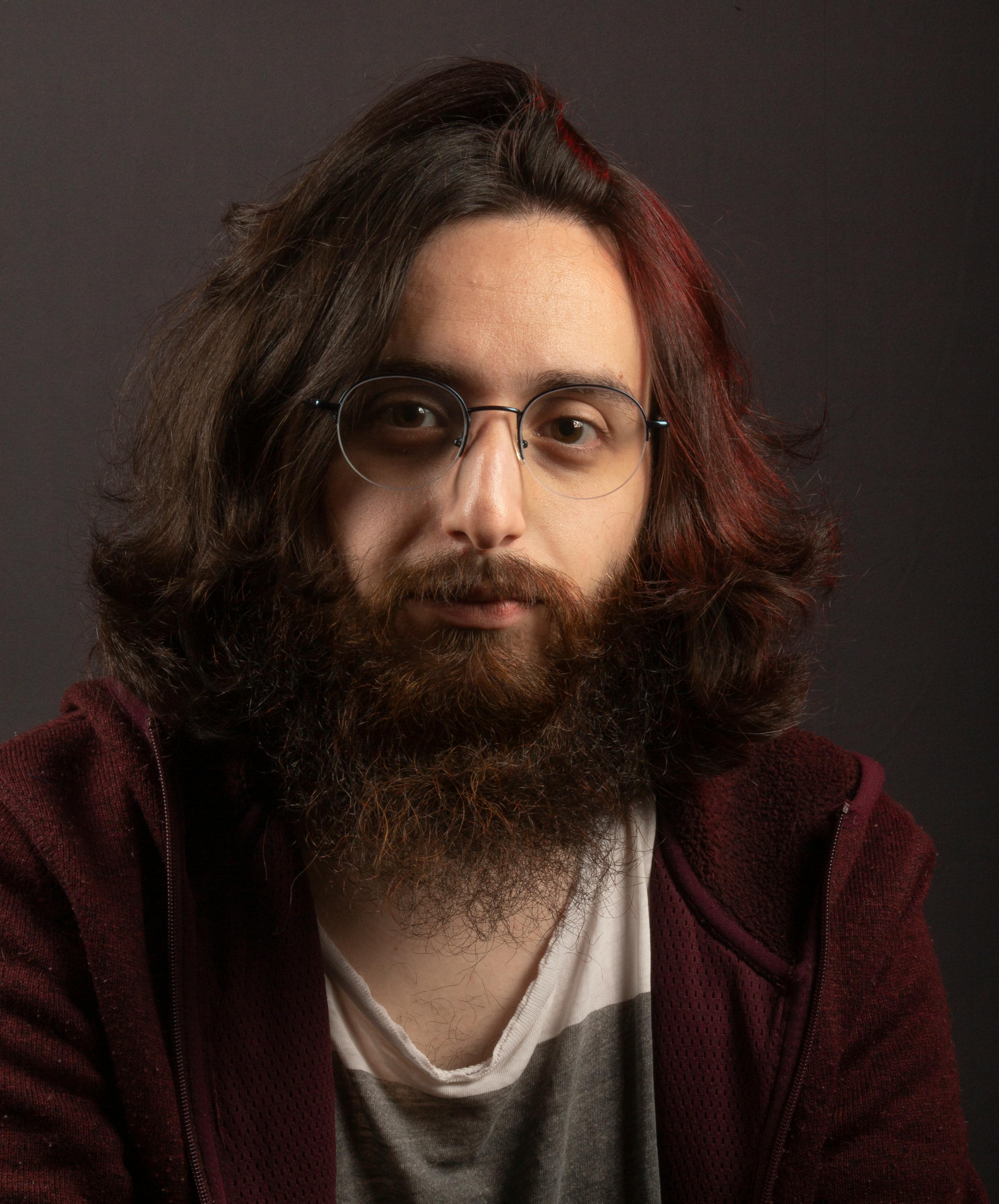 Close-up portrait of a young bearded man wearing glasses, neutral expression.