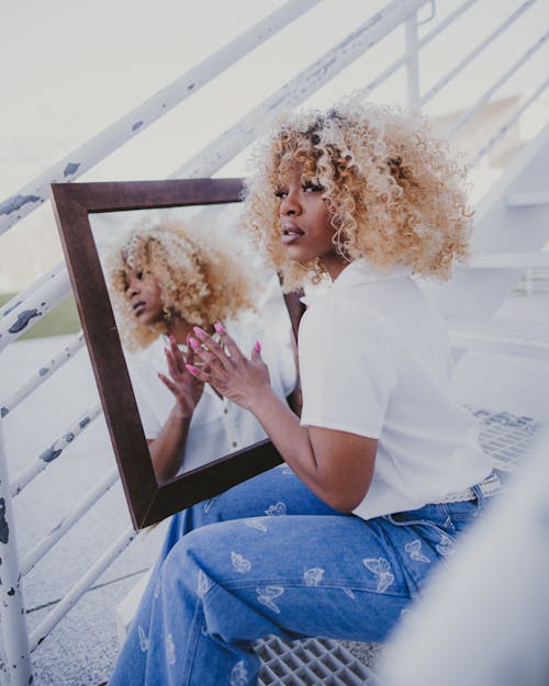 Curly-Haired Woman in White Shirt Holding a Mirror while Sitting on Staircase
