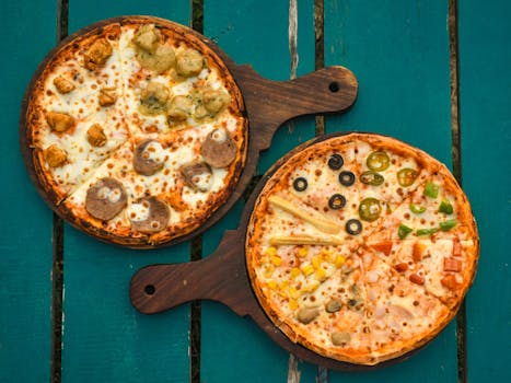 Two pizzas with various toppings on wooden boards over a rustic green table, captured in a top-down view.