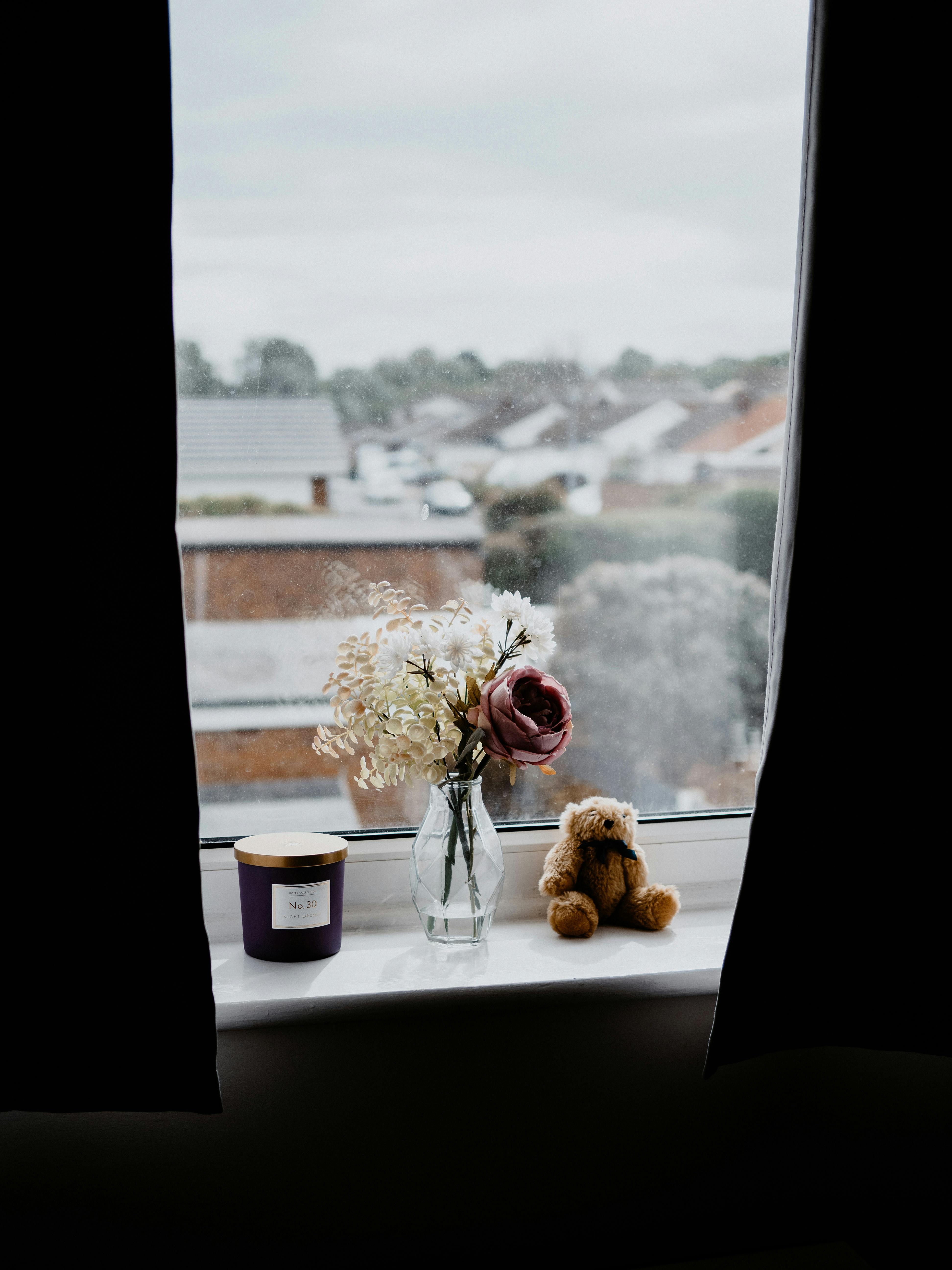 Intimate view of a windowsill with a flower vase, candle, and teddy bear, overlooking suburban homes.