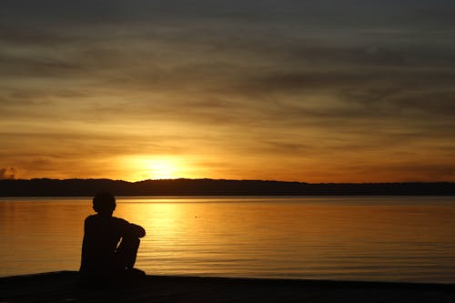 Silhouette of a Man Sitting Alone on Sea Shore during Sunrise