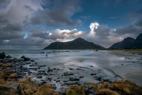 Free Scenic view of a rocky shore with dramatic clouds, mountains, and a person by the sea on a cloudy day. Stock Photo