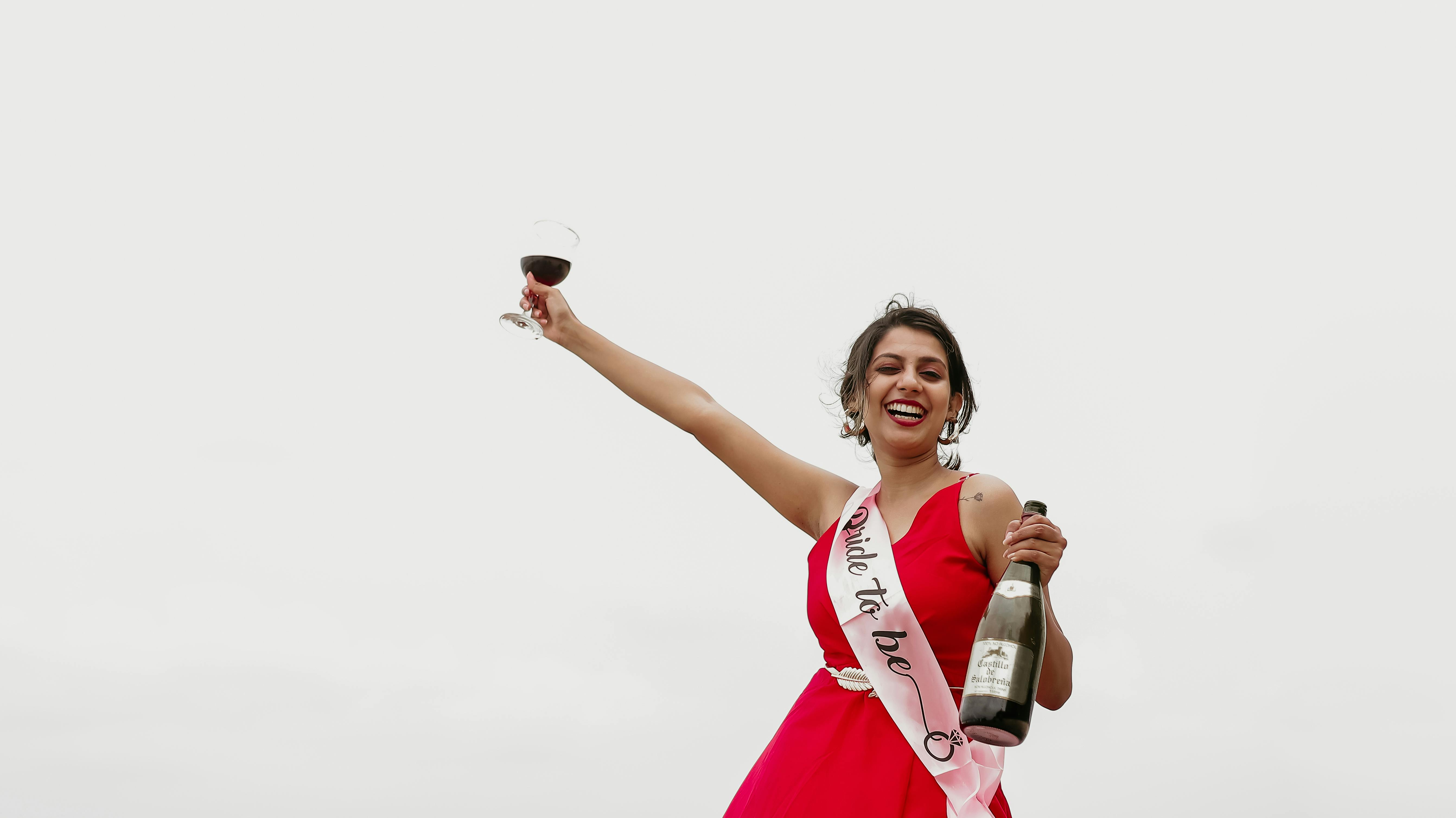A cheerful bride-to-be in a red dress celebrates with wine outdoors.