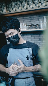 Barista in apron and face mask holding a cup in a cafe setting, symbolizing new normal