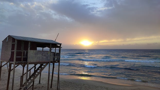Tranquil sunrise over a sandy beach with a rustic lifeguard hut silhouetted against the calm waves.