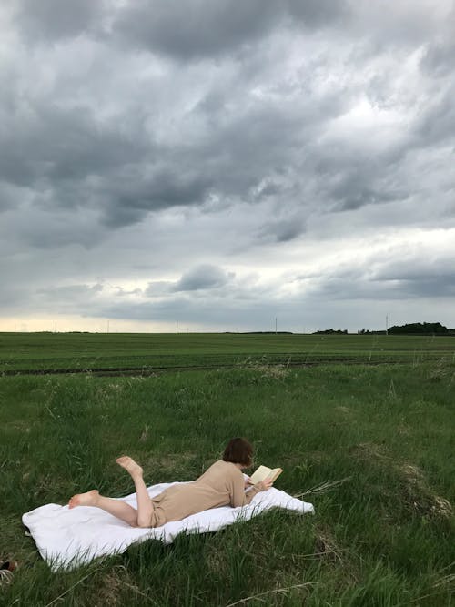 Woman Lying on a White Blanket on the Middle of the Grass Field while Reading a Book Under Cloudy Sky