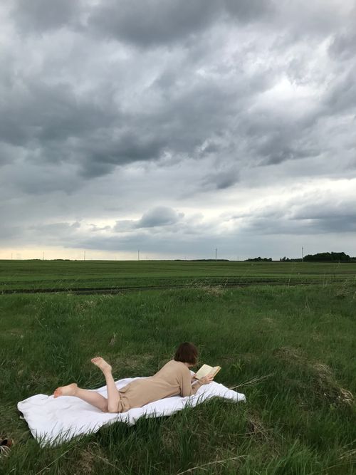 Free A woman reads a book while lying on a blanket in a grass field under a cloudy sky. Stock Photo