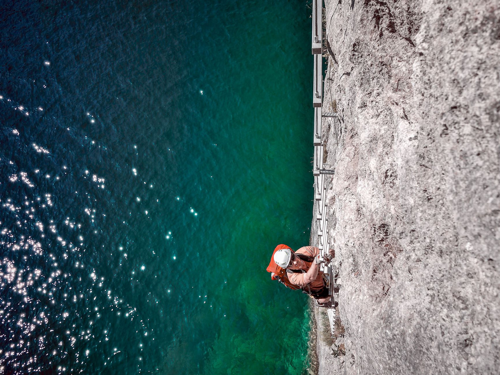 A rock climber ascends a cliffside overlooking sparkling blue-green water.
