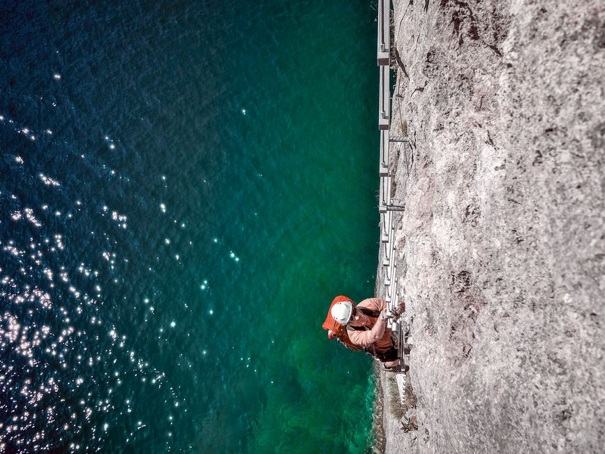 A rock climber ascends a cliffside overlooking sparkling blue-green water.