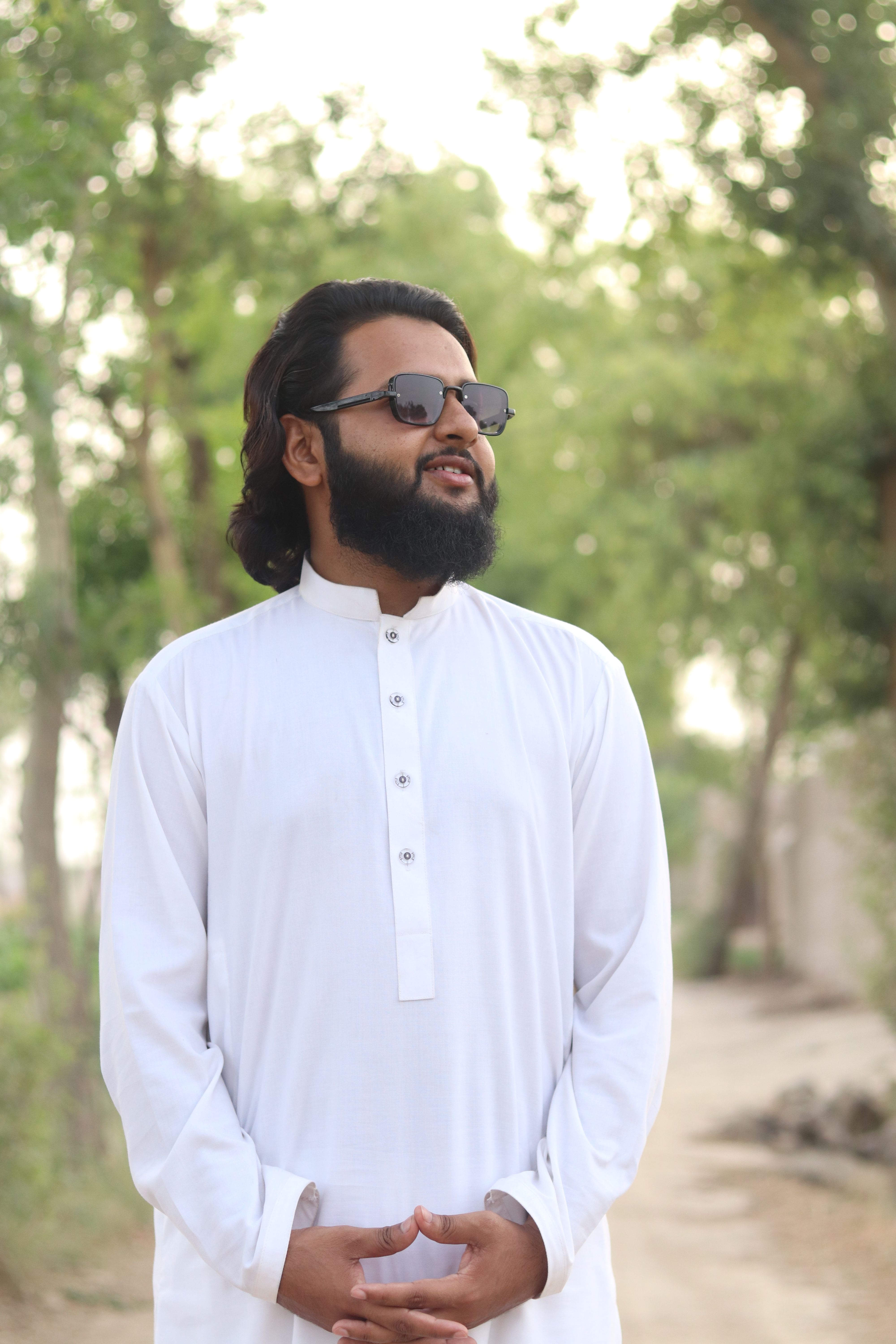 Bearded man in a white kurta and sunglasses stands confidently outdoors against a leafy backdrop.