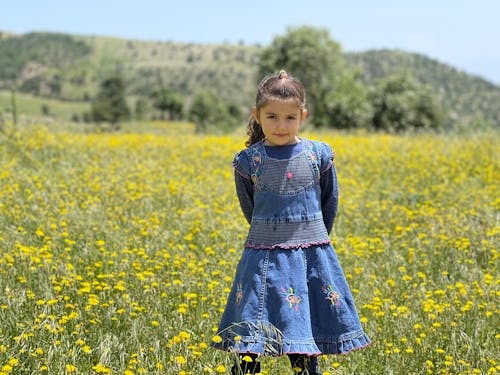 A Cute Little Girl in Denim Dress Standing on a Flower Field while Smiling at the Camera