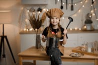 Little Girl Dressed as a Chef Sitting on a Kitchen Counter Holding a Pot and a Soup Spoon 