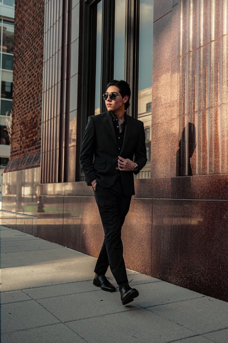 A Man In Black Suit Walking On The Sidewalk