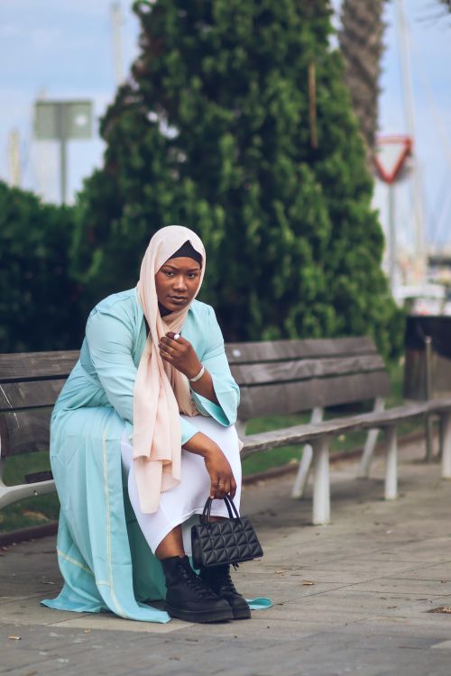 Free Stylish woman wearing hijab sits on a wooden bench in a tranquil park setting, exuding elegance and fashion sense. Stock Photo