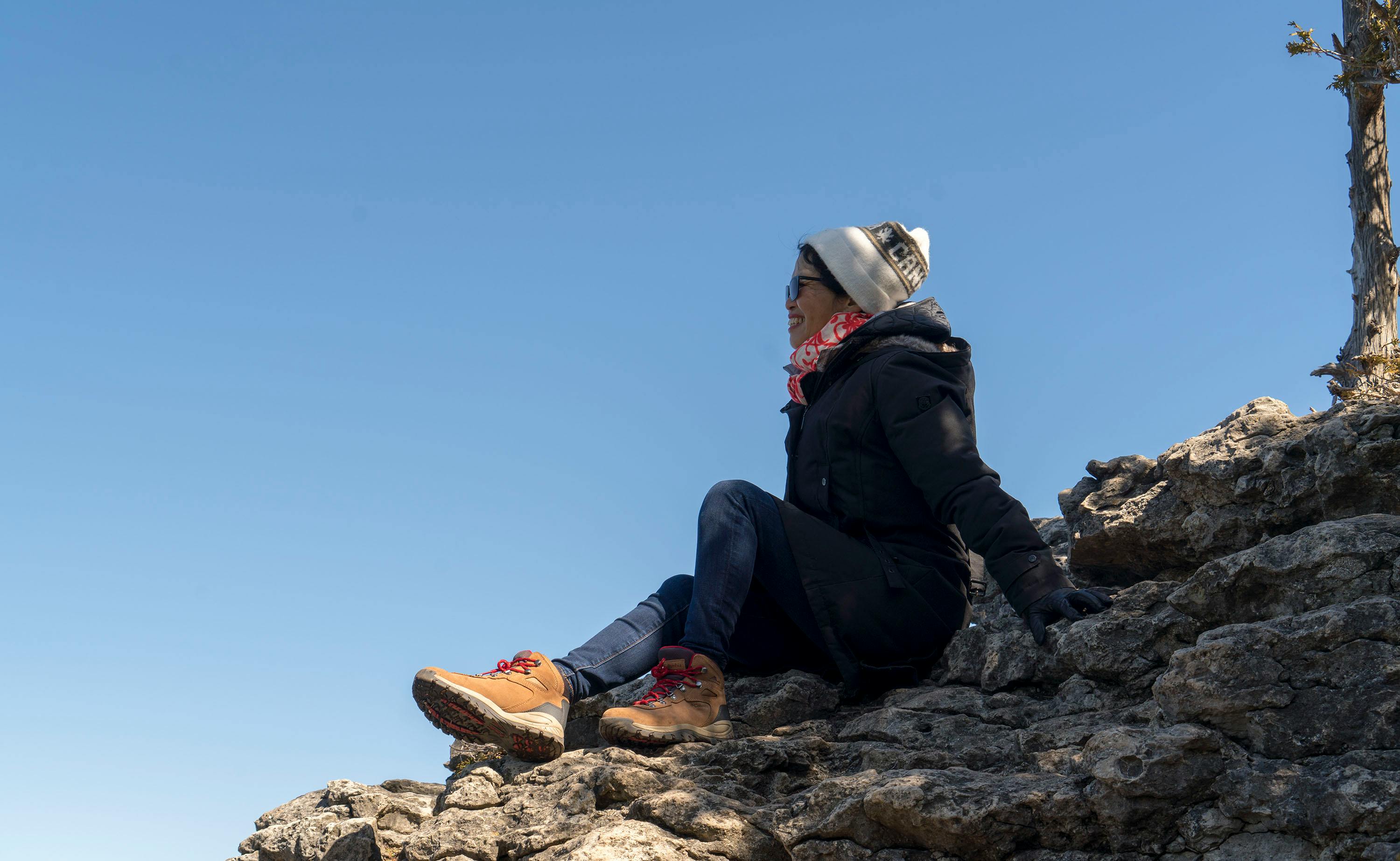 Woman sits on rocky terrain, enjoying a sunny day outdoors, dressed warmly for adventure.