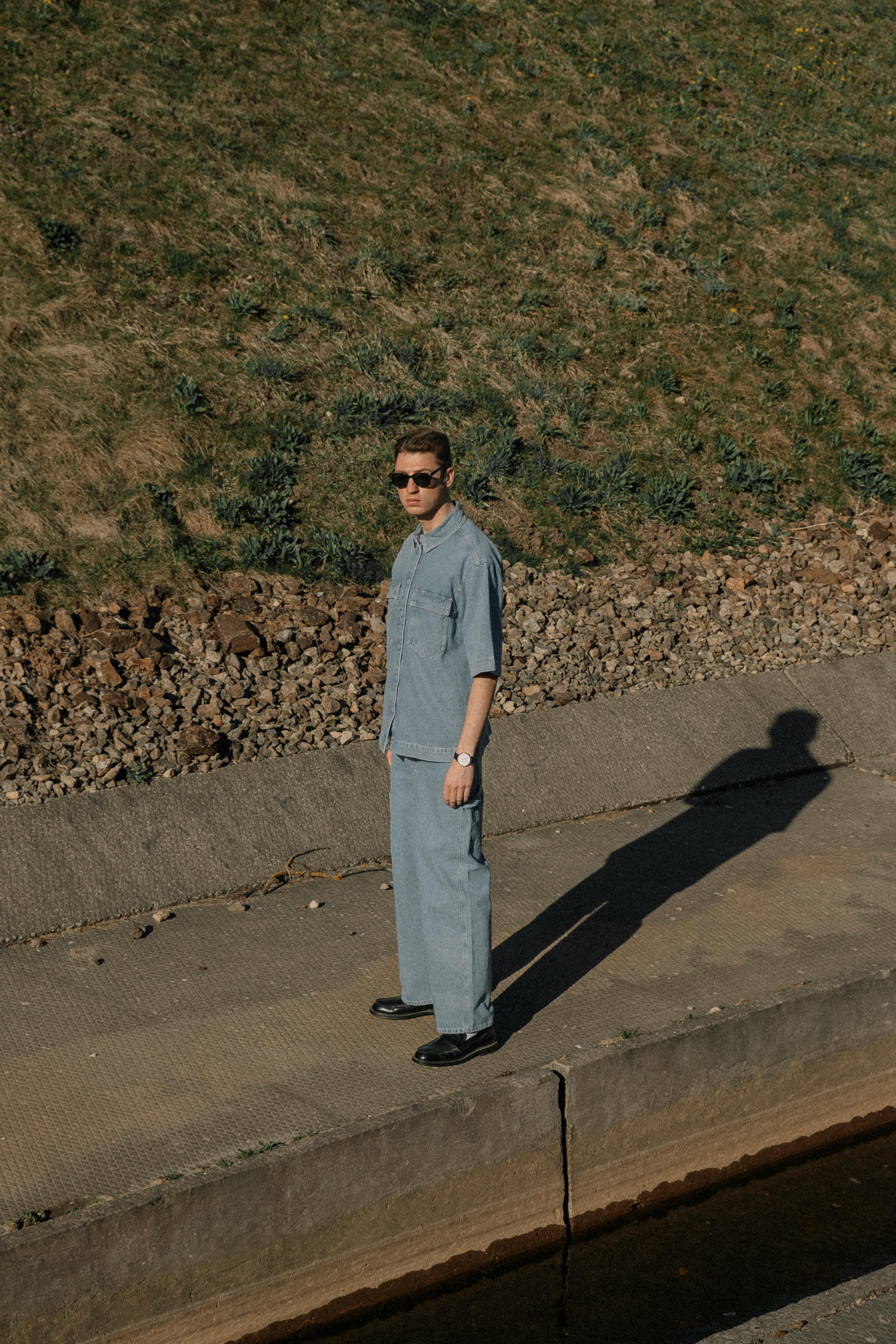 Stylish man in blue attire with sunglasses posing outdoors under natural light.
