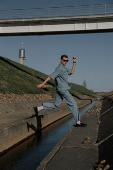 Energetic young man in denim jumping near a canal under a bridge, showcasing urban street style.