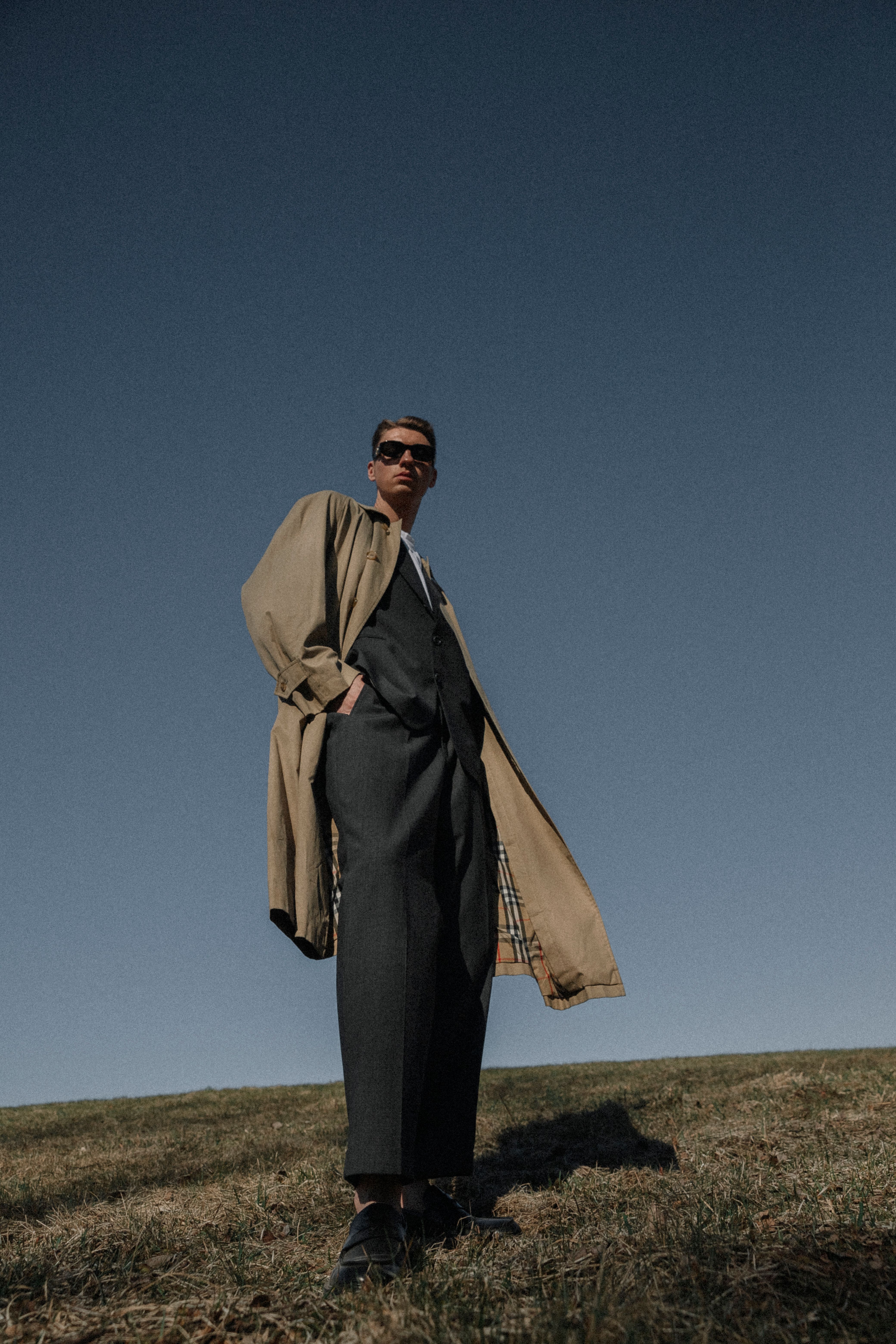Man standing on grassy field wearing a stylish coat under a clear blue sky.
