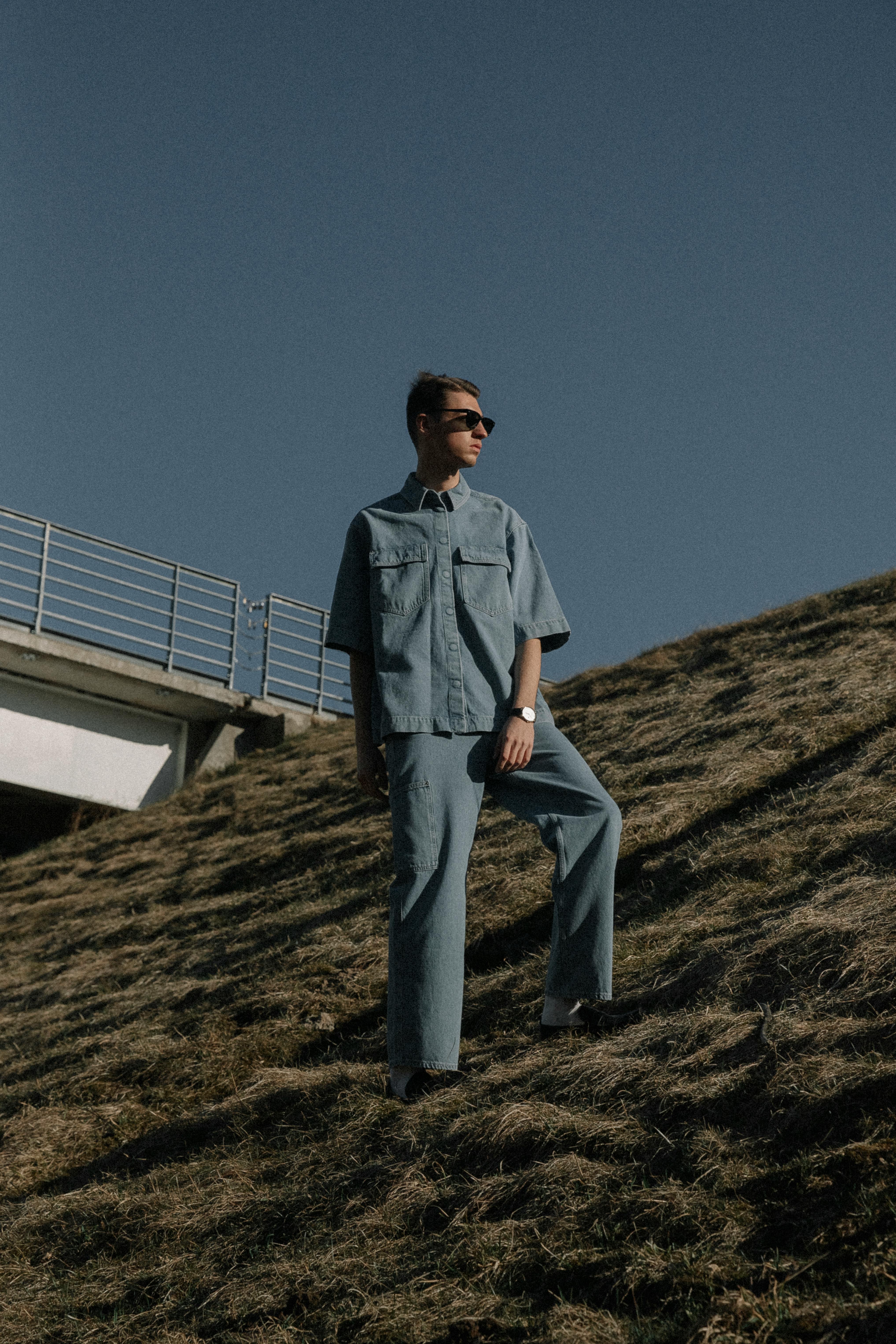 Stylish man wearing denim attire posing on a grassy slope with a clear blue sky.